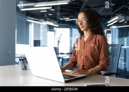 Young beautiful businesswoman working inside modern office building, arab woman with curly hair and glasses using laptop, smiling and happy Stock Photo