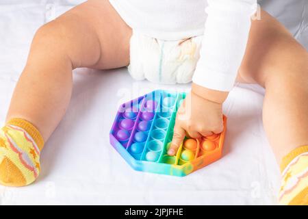 Newborn baby sits on the bed playing trendy flexible silicone multicolored toy pop it. Selective focus on anti stress simple dimple toy in children's Stock Photo