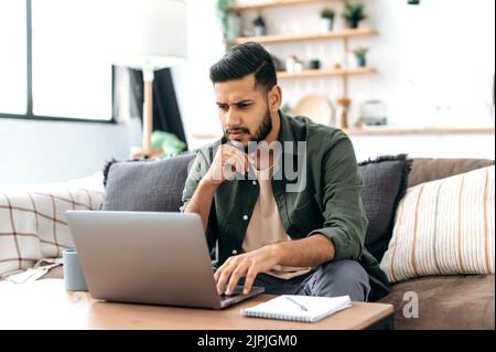 Focused smart arabian or indian young man in stylish casual clothes, freelancer, web designer, sitting in the living room on the sofa, working on a laptop on a project, looking at the screen intently Stock Photo
