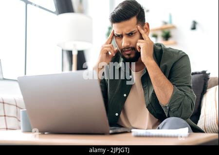 Tense focused tired arabian or indian stylish guy, freelancer, sitting on a sofa in a living room, using laptop for work or study remotely, massaging his temples, feeling overworked, headache Stock Photo