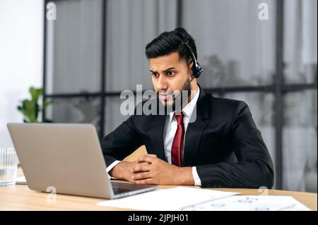 Puzzled stunned indian or arabian young businessman, company ceo, with headset, in a suit, look at laptop screen in confusion, see unexpected message or news, surprised emotional facial expression Stock Photo