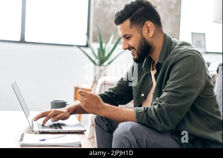 Side view of satisfied positive indian or arabian guy in stylish clothes sitting on a sofa in a living room, using laptop and bank card to pay online, ordering goods and clothes, home delivery, smiles Stock Photo