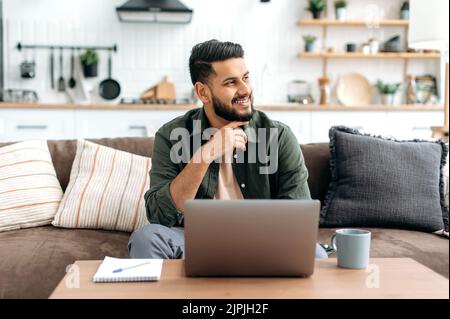 Handsome confident thoughtful arabian or indian guy in casual wear, freelancer, sit on sofa at home in living room, using a laptop for work, looking pensively away, dreaming, thinking, smiling Stock Photo