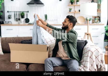 Excited arabian or indian man in casual clothes, sitting on a sofa in the living room, unpacking his parcel, taking out clothes from the cardboard box, looking at her happily, satisfied with new wear Stock Photo