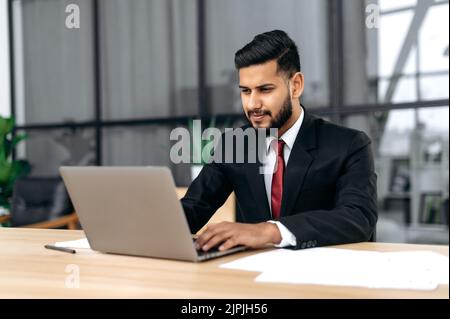 Focused intelligent confident arabian or indian successful businessman, company ceo, in formal suit, sit at work desk with laptop in modern creative office, concentrated looks at laptop screen Stock Photo