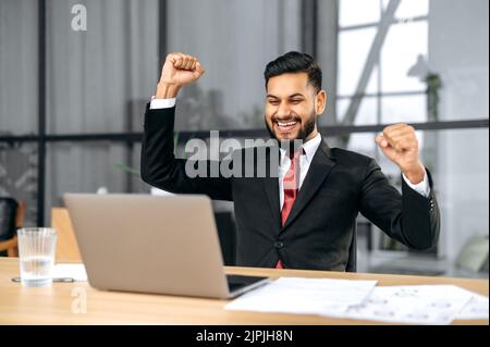 Excited indian or arabian businessman in suit, sits in a modern office, looking at laptop, happy by good news, rejoice by win, raising hand in yes gesture, celebrating project success, big profit Stock Photo