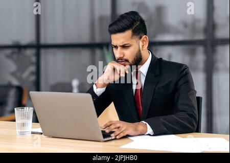 Confused puzzled indian or arabian young businessman, entrepreneur, in a suit, sitting in his modern office, focused look at laptop screen, see unexpected message or news, is under stress Stock Photo