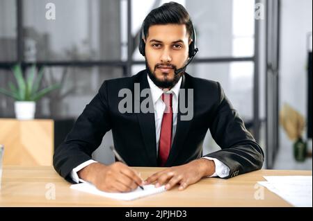 Photo of successful serious indian or arabian young businessman, financial mentor, with headset, sit at the desktop in his modern office, looking at the camera, listening interlocutor, takes notes Stock Photo
