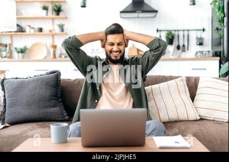 Shocked confused indian or arabian young man, freelancer, designer, looking surprised at laptop screen, stressed from the failure of a deal or project, read unexpected news, in desperation, annoyed Stock Photo