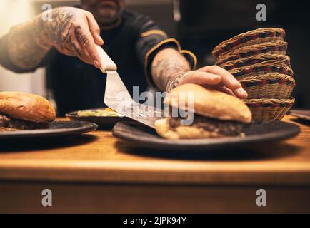 Order up. Closeup of a unrecognisable chefs tattooed hands serving up a burger into a plate inside of a kitchen of a restaurant. Stock Photo