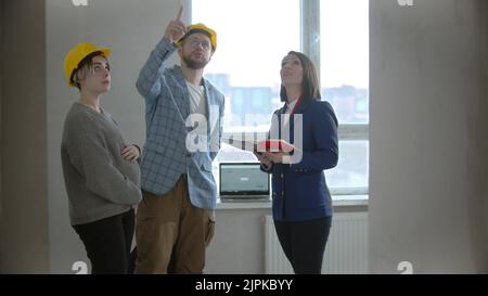 A pregnant woman with her husband talking with a real estate agent in apartment in a new building - man asking questions and pointing at the ceiling Stock Photo