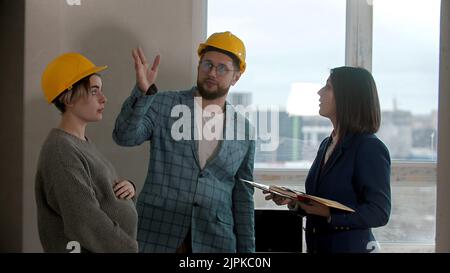 A pregnant woman with her husband talking with a real estate agent in apartment in a new building - man asking questions and pointing at the walls Stock Photo