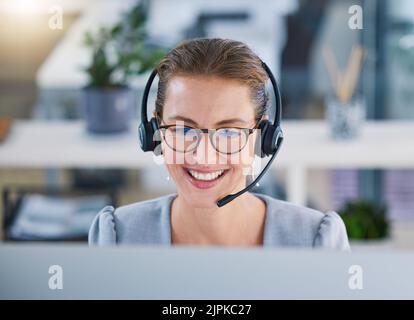Call center agent helping clients online while talking on a headset in a modern office, enjoying her career. Customer service and support with a happy Stock Photo