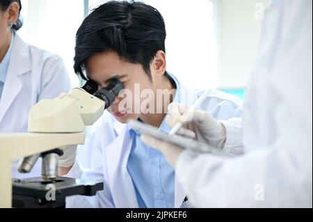 Talented young Asian male scientist looking under the Microscope, working with his colleagues in the laboratory. Pharmaceutical research concept Stock Photo