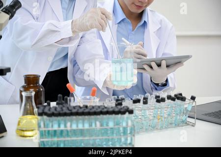 A professional female scientist adjusts a chemical liquid in the beaker while a male scientist records the result on the tablet. cropped image Stock Photo