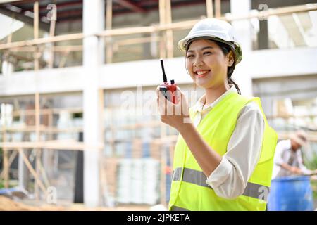 Attractive young Asian female engineer or architect in white safety helmet using a walkie talkie in the construction site. Stock Photo