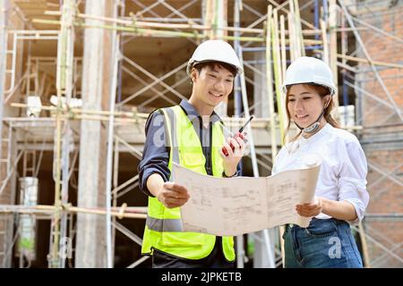 A professional Asian male engineer and attractive Asian female architect are looking and holding a blueprint in front of the construction site. Stock Photo