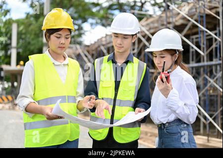 Two talented young Asian female and male architects working and inspecting the building with female construction inspector at the construction site. Stock Photo