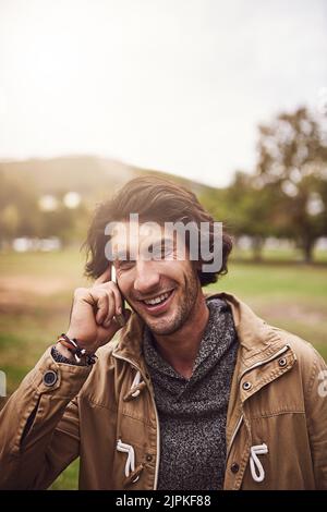We can talk for hours. a cheerful young man talking on his phone while standing outside in a park. Stock Photo