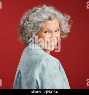 .. Confident, happy and smiling senior woman feeling playful and cheerful while posing against red studio background. Portrait of a beautiful, older Stock Photo