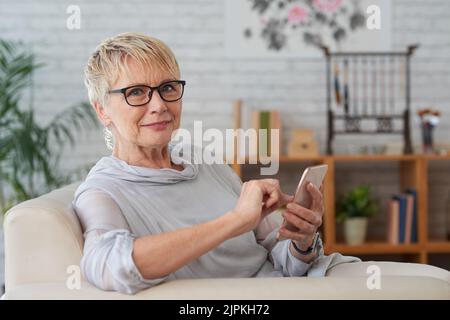 Elderly womn sitting on sofa and using application in her smartphone Stock Photo