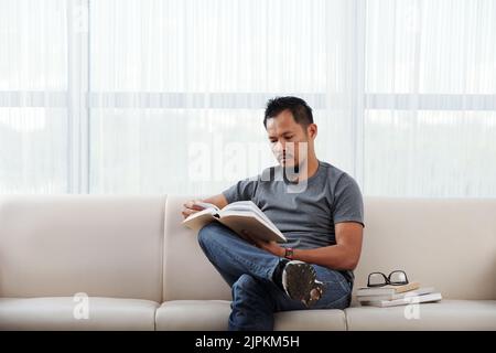 Filipino man sitting on sofa in his living room and reading a book Stock Photo