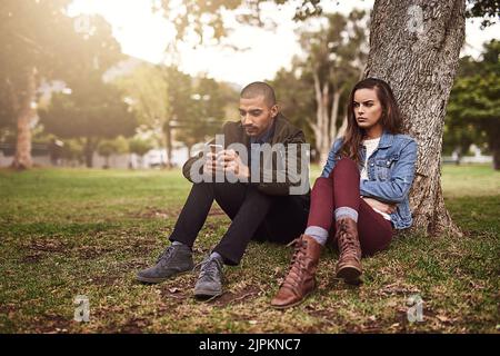 He gives that phone too much attention. a cheerful young man sitting down under a tree with his unhappy girlfriend while using his phone outside in a Stock Photo