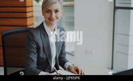 Portrait of Attractive blonde businesswoman sitting at table writing in notebook smiling into camera in modern office indoors Stock Photo