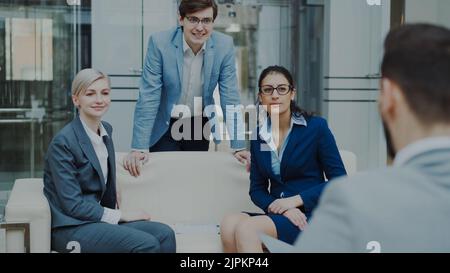 Team of cheerful business colleagues talking and discussing each other while having break sitting on sofa in modern office hall indoors Stock Photo