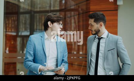 Two businessmen colleagues walking and discussing documents in modern office hall indoors Stock Photo