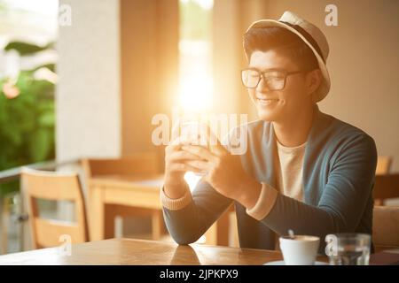 Cheerful Asian young man sitting in sunny cafe and reading text messages in his smartphone Stock Photo