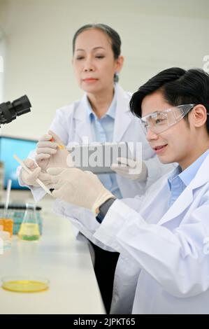 Professional Asian-aged female scientist instructs her young male scientist to adjust a new chemical liquid in the lab. Stock Photo