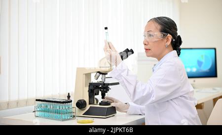 Successful and professional Asian senior female scientist supervisor working in the laboratory, analyzing the liquid sample, using a microscope. Stock Photo