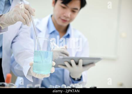 Handsome young Asian male scientist working in the lab, recording the experiment results on tablet while his coworker adjusting a chemical liquid in a Stock Photo