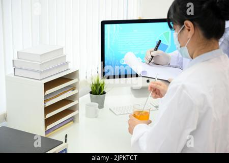 Concentrated young Asian female scientist or chemist working with her team in the lab, adjusting a chemical liquid in a beaker and recording the exper Stock Photo