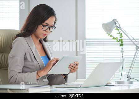 Young Asian business woman in glasses reading information on digital tablet Stock Photo