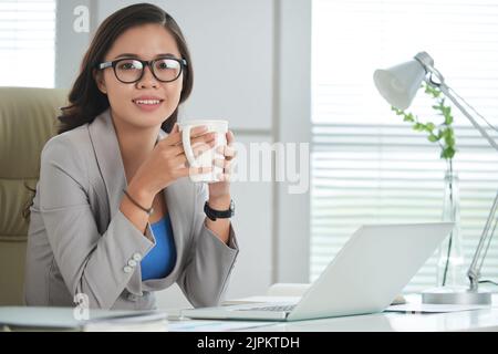 Portrait of pretty Vietnamese business lady with big mug of coffee sitting at her table Stock Photo