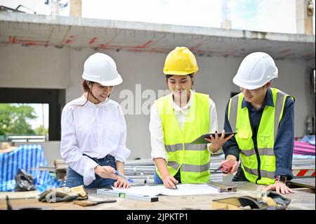 Attractive Asian female architect checking some details on blueprint during meeting with an engineer and construction inspector at the construction si Stock Photo