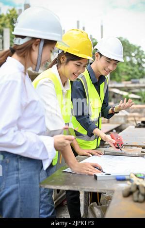Team of a cheerful and professional millennial Asian civil engineer checking the blueprint together at the construction site. Stock Photo