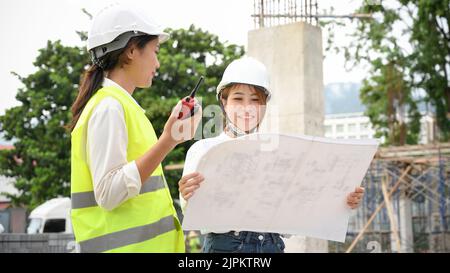 Two talented young Asian female civil engineer or architect discussing, looking at the blueprint and working together at the construction site. Stock Photo