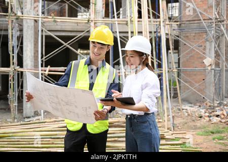 Beautiful and talented young Asian female civil engineer looking at the blueprint during discussing with a professional male construction inspector at Stock Photo
