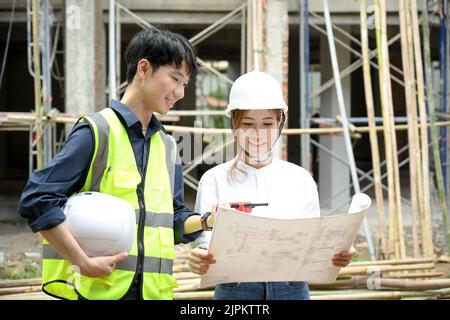 Attractive young Asian female architect looking at the blueprint while talking with professional male engineer in the construction site. Stock Photo