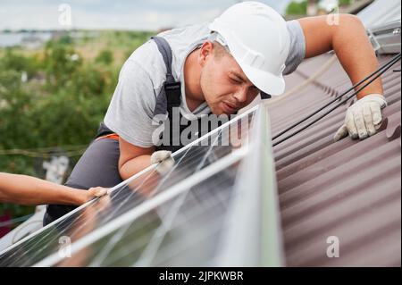 Men mounters mounting photovoltaic solar moduls on roof of house. Close up electrician in helmet looking on evenness of solar panel during installing. Concept of alternative and renewable energy. Stock Photo
