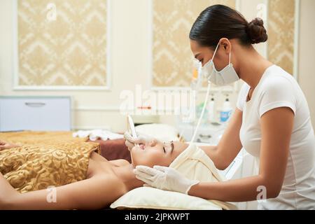 Profile view of pretty young woman lying on treatment table of beauty salon while getting ultrasound face cleaning Stock Photo