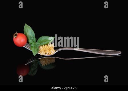 A large metal fork with pasta, basil and a tomato on it. The cutlery is reflected against a dark background. Stock Photo