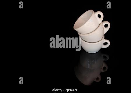 Three white, old espresso cups are stacked on the edge. The background is black. The cups are reflected on the ground. There is space for text. Stock Photo