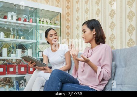 Beautiful Asian cosmetologist asking her female client about desired result from treatment and taking necessary notes on digital tablet while sitting at modern facial room Stock Photo