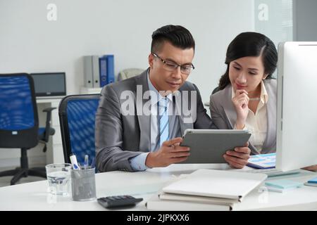 Pensive Vietnamese business people discussing information in tablet computer Stock Photo