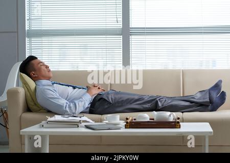 Tired Vietnamese businessman sleeping on sofa in his office Stock Photo