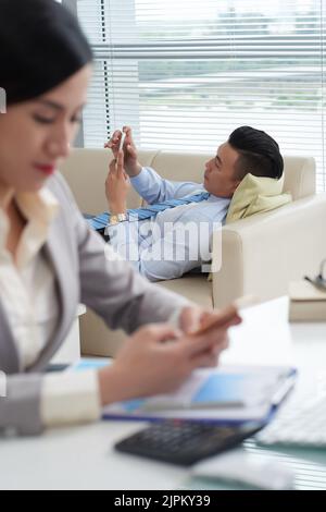Vietnamese businessman lying on sofa and using application on his smartphone Stock Photo